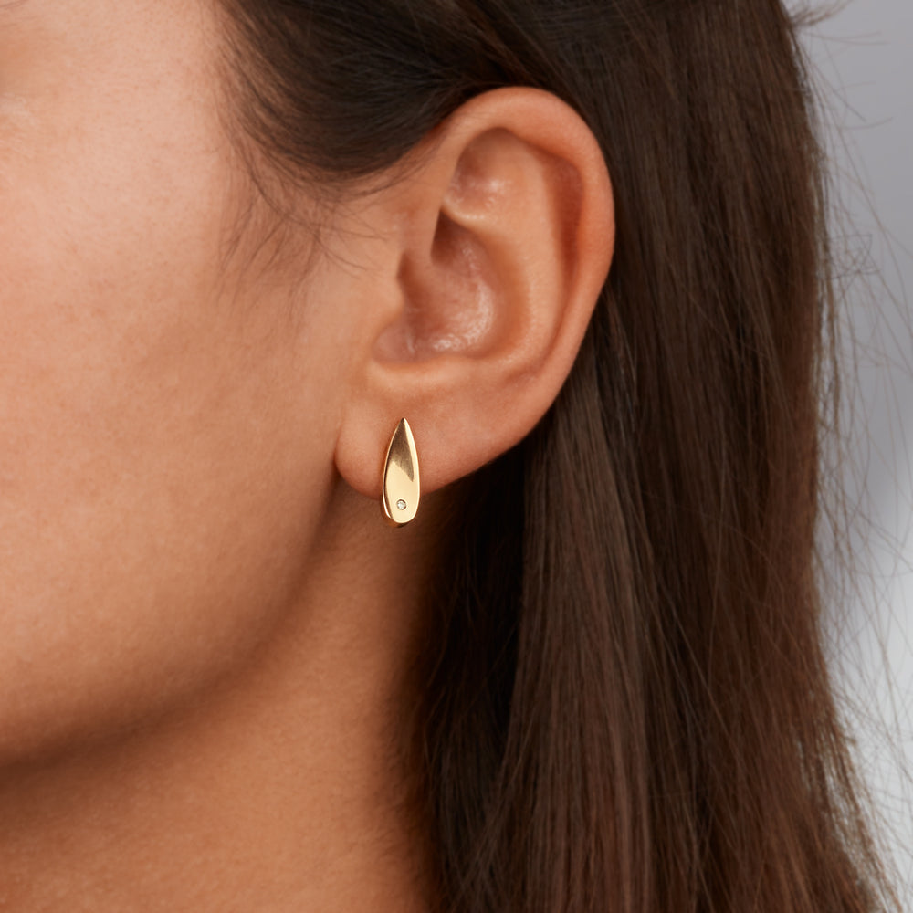 Een close-up toont het oor van een persoon met elegante Boccia Titanium Oorstekers gp dia 2/0.01 - 05071-06. Deze oorstekers hebben een gouden druppelvorm met een delicate edelsteen tegen een achtergrond van gedeeltelijk zichtbaar donker haar en een neutrale, wazige achtergrond.