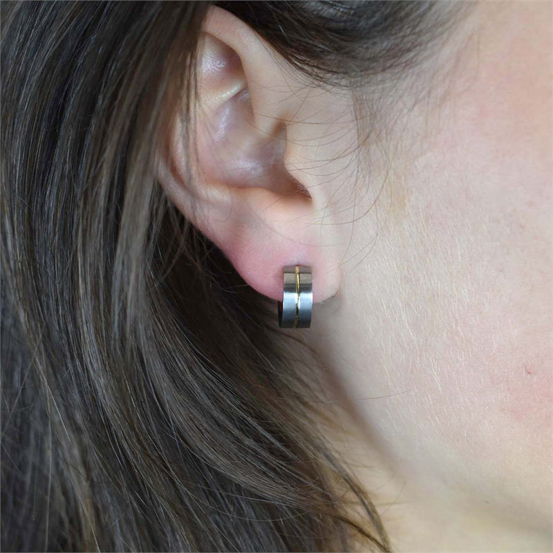 Close-up van het oor van een persoon met Boccia Titanium Oorringen Bicolor 0503-03, kleine dikke zilveren oorringen met een zwarte streep. Het bruine haar bedekt het oor gedeeltelijk, wat contrasteert met deze titanium oorbellen van Boccia.