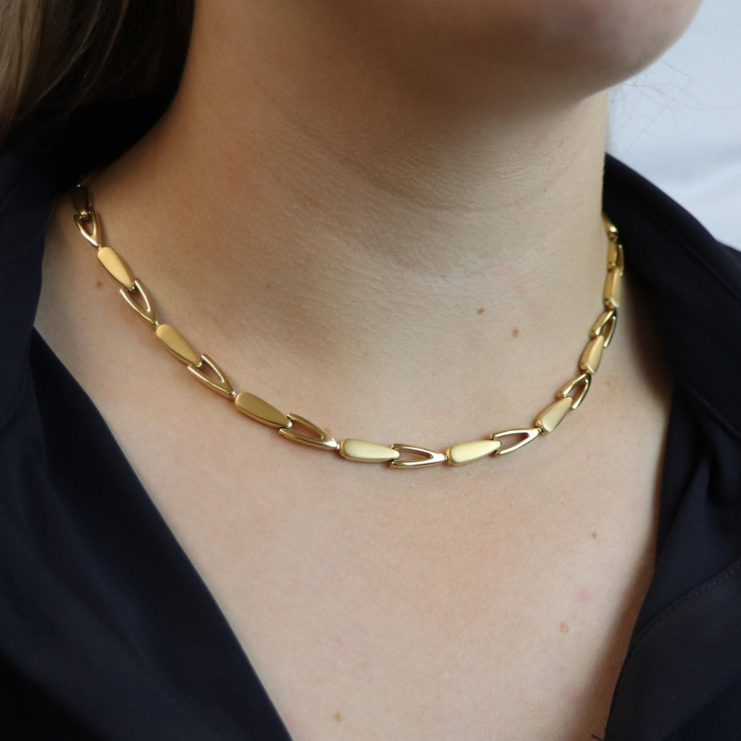 Een close-up van een persoon die de Boccia Titanium Collier gp 08039-03 draagt, een goudkleurige ketting met grote platte schakels van Boccia, gecombineerd met een donkere top. Alleen de hals en kraag zijn zichtbaar, wat deze stijlvolle sieraden extra benadrukt.