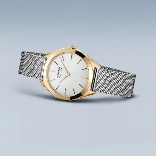 De Bering 17331-010 dameshorloge heeft een gouden kast, witte wijzerplaat met minimale markeringen en een zilverkleurige mesh band. Het saffierglas zorgt voor duurzaamheid terwijl het in een lichte hoek tegen een lichtblauwe achtergrond rust.