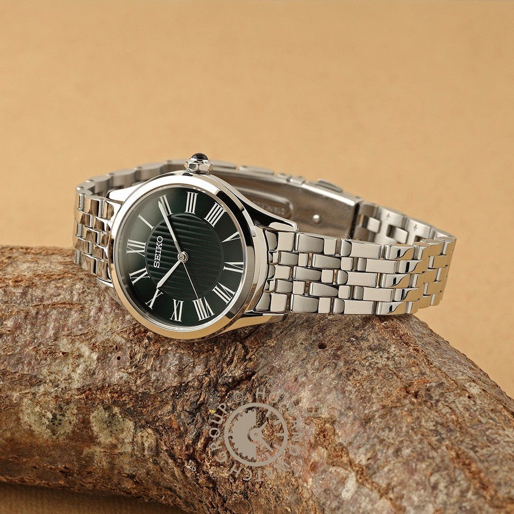 Een zilveren Seiko dameshorloge SUR611P1 met een metalen band, zwarte wijzerplaat, saffierglas en Romeinse cijfer uurmarkeringen rust op een gestructureerde bruine tak tegen een bruine achtergrond.