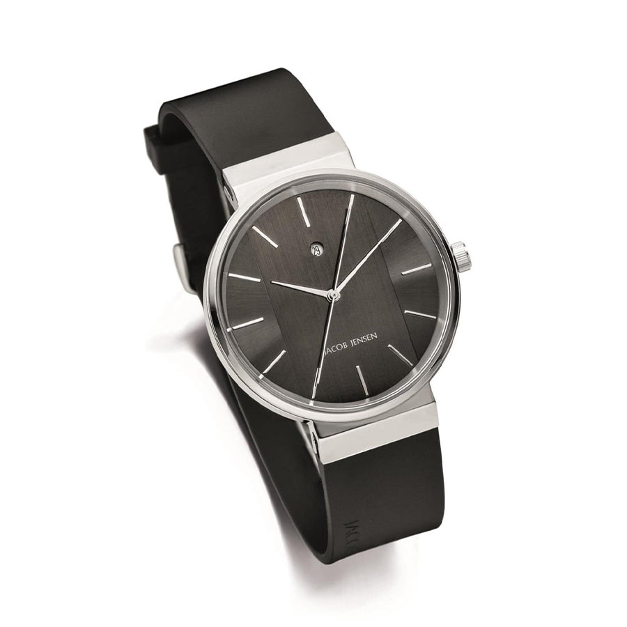 Maak kennis met de Jacob Jensen 708, een strak analoog horloge met een zwarte wijzerplaat, minimalistische zilveren uurmarkeringen en een enkele diamant. Het heeft een zwarte rubberen band en zilveren lunette, wat moderne verfijning belichaamt.