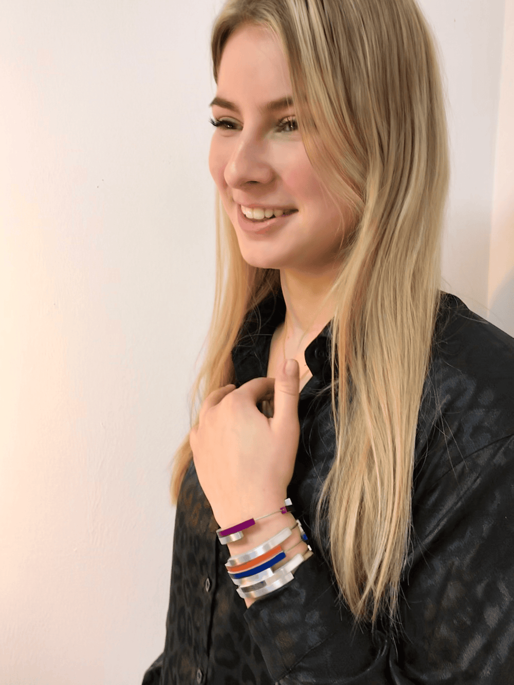 Een persoon met lang blond haar lacht terwijl hij een zwart gebloemd shirt en verschillende kleurrijke armbanden laat zien, waaronder de paarse Armband Vier bogen Paars A32P van CLIC Creations, handgemaakt in Nederland. De effen witte muur dient als achtergrond.