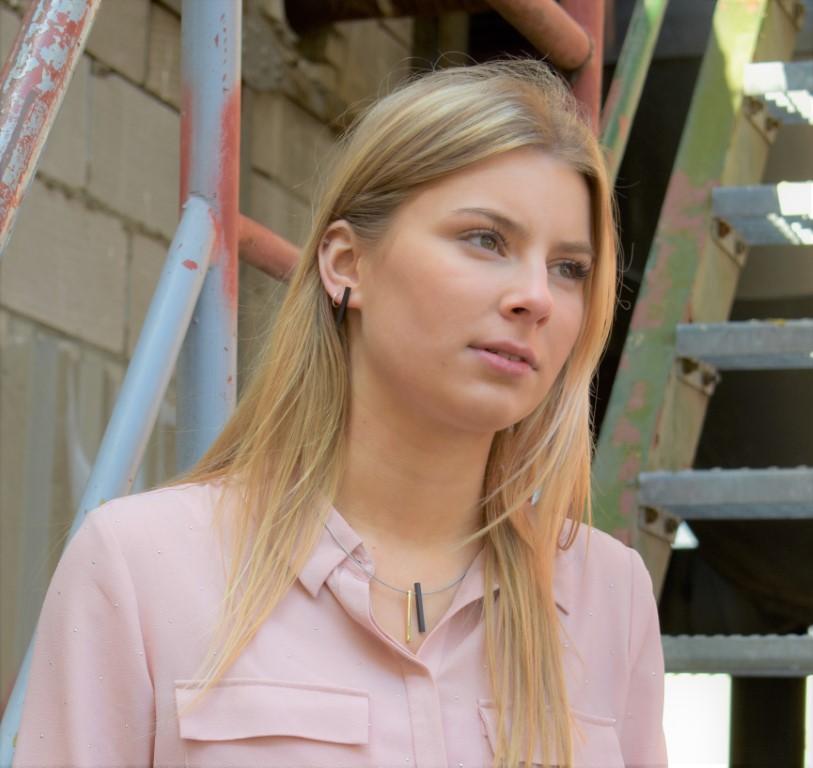 Voor een metalen trap en een bakstenen muur draagt een vrouw met lang blond haar een lichtroze blouse en toont ze de Oorsteker Staafje Goud|Geel O26G van CLIC Creations. Ze draagt de hypoallergene aluminium ketting en kijkt met een neutrale uitdrukking iets weg van de camera.