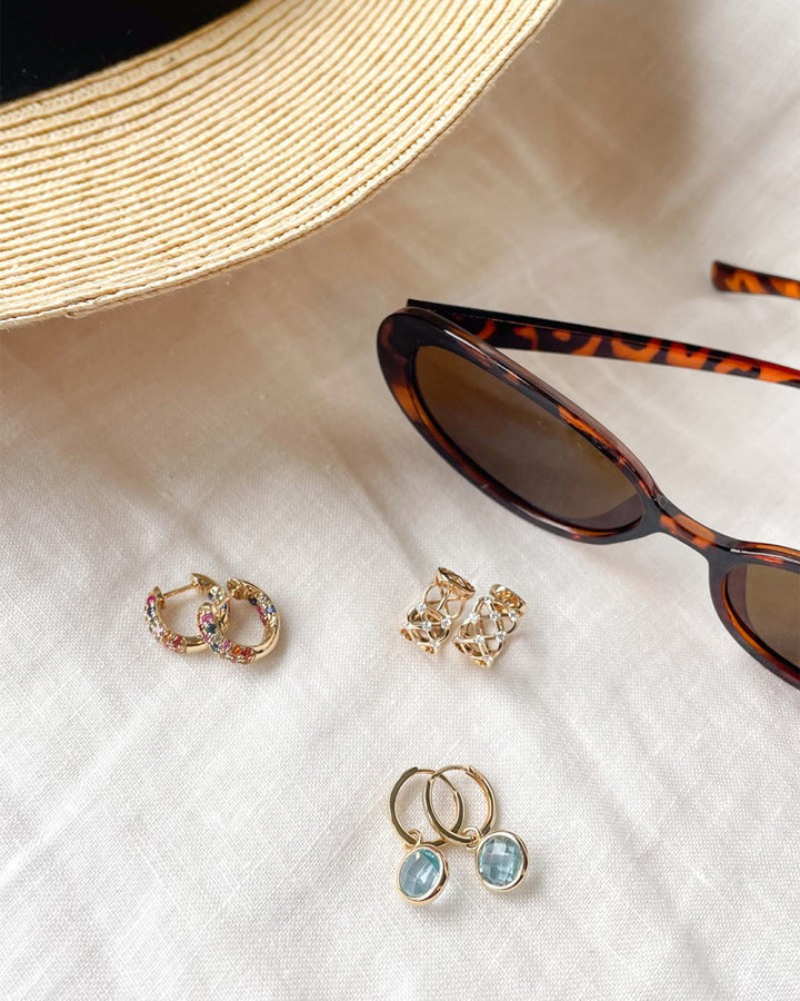 Een beige zonnehoed, een zonnebril met schildpad en drie paar oorbellen, waaronder de Geelgouden Oorringen met Regenboog Saffier in Luxe Pavézetting van Excellent Jewelry, zijn kunstig gerangschikt op een lichte stoffen achtergrond.