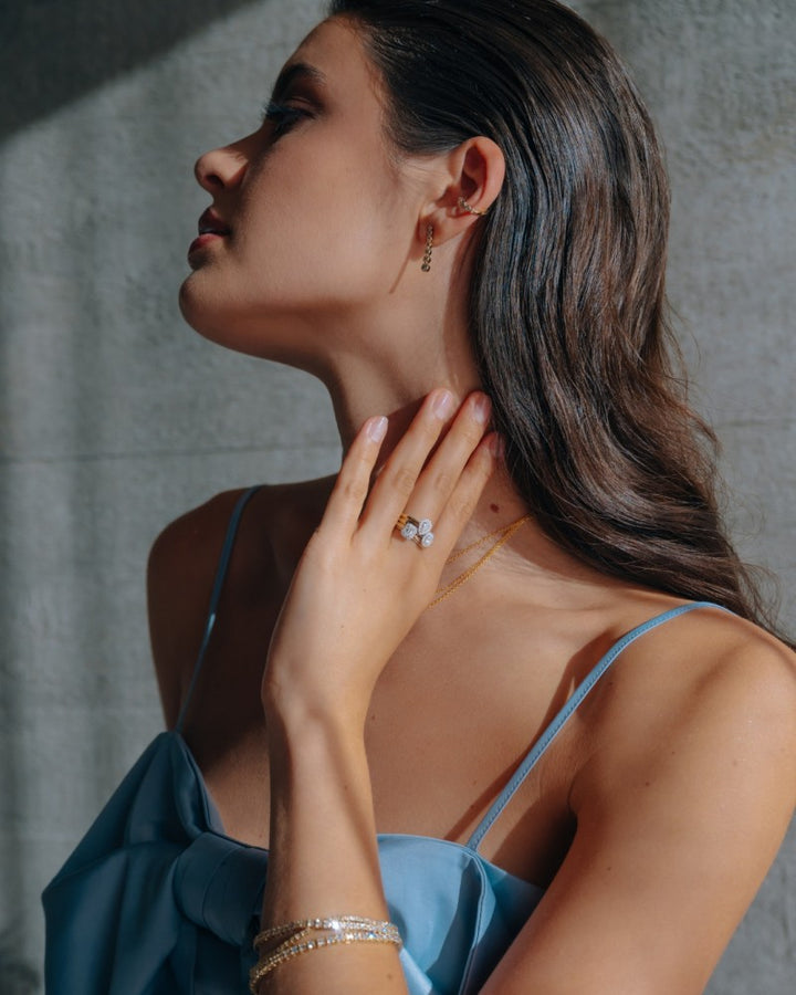 Een vrouw in een lichtblauwe jurk met spaghettibandjes raakt haar hals aan en toont de 14k Bicolor Gouden Ring van Excellent Jewelry met een ovaal diamanten hart in entourage zetting, samen met bijpassende gouden halsketting, armband en oorbellen tegen een zonovergoten grijze achtergrond.