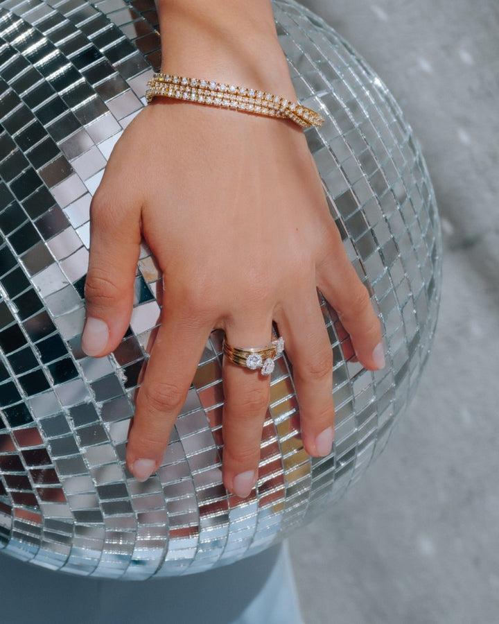 Een hand die de 14k Bicolor Gouden Ring van Excellent Jewelry draagt met een smaragd geslepen diamant in een entourage zetting rust op een reflecterende discobal, zachte verlichting accentueert de schittering van de ring.
