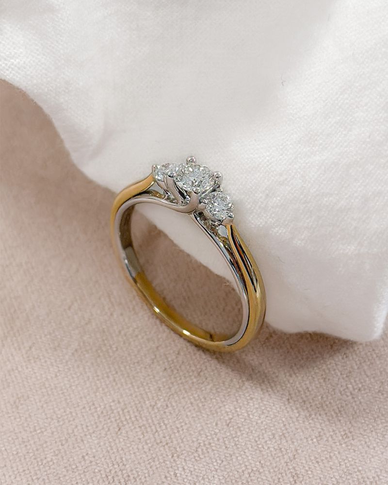 De Bicolor Geel- en Witgouden Damesring met Diamant (0.51 crt) van Excellent Jewelry, 14 karaat, schittert met drie ronde diamanten bovenop en ligt elegant op een zachte witte stof.