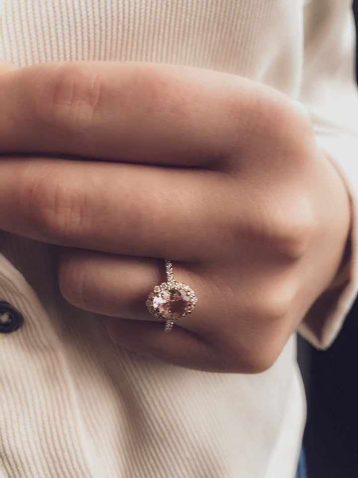 Een hand draagt de Elegante Geelgouden Ring met Roze Morganiet en Briljante Diamanten van Excellent Jewelry, met een grote ovale edelsteen omringd door diamanten. De persoon heeft een crèmekleurig geribd overhemd aan met donkere knopen.