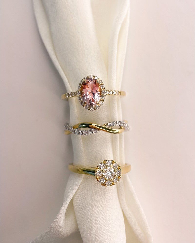 Drie elegante Geelgouden ringen van Excellent Jewelry liggen op witte stof: de bovenste met roze morganiet en briljante diamanten, de middelste met een gedraaid diamantenmotief en de onderste met een cluster van kleine diamanten.