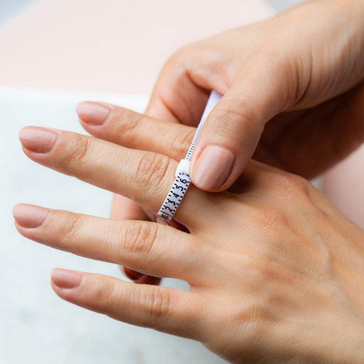 Een persoon gebruikt de Pas ring voor de perfecte ring maat van Juwelier van der Kooij om zijn of haar perfecte ringmaat te vinden, waarbij zijn of haar verzorgde nagels afsteken tegen een zacht wazige achtergrond.