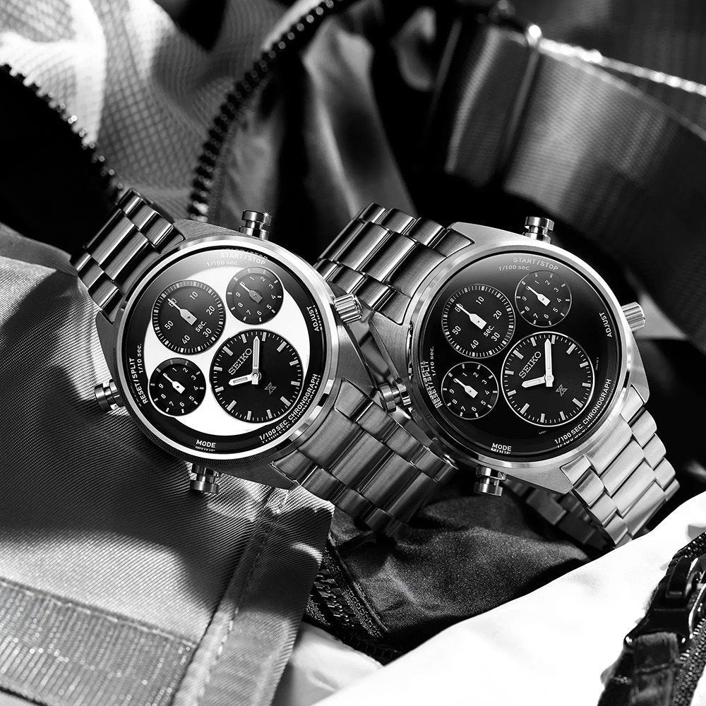 Twee Seiko Prospex SFJ003P1 chronograafhorloges van roestvrij staal, met zwarte wijzerplaten en subdials, liggen op een gestructureerde stof. Eén heeft een contrasterende witte wijzerplaat. Beide hebben schakelarmbanden, die het klassieke Seiko-ontwerp laten zien.