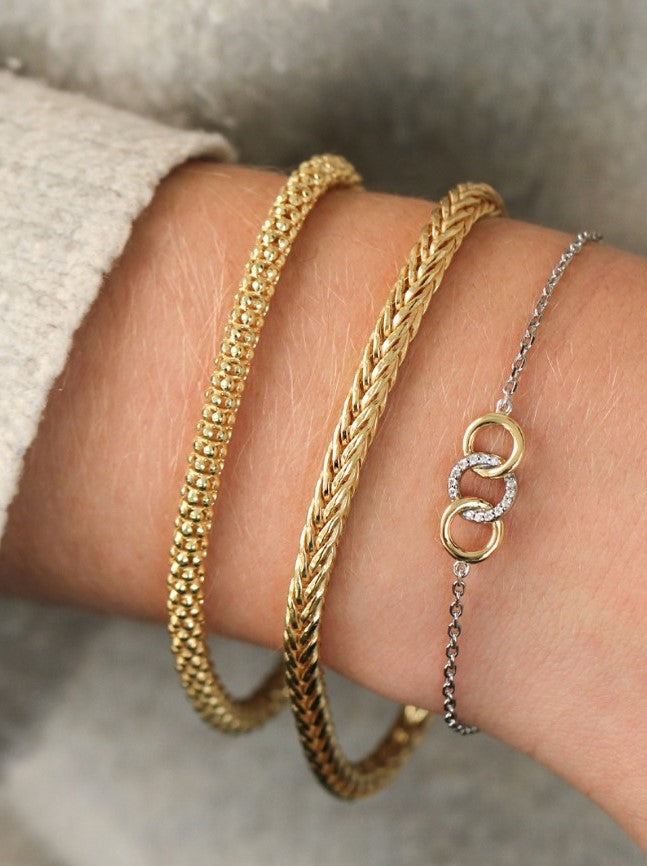 Een close-up van een pols toont de elegante Zinzi Armband Gold Plated Zia1925 G: twee gouden kettingarmbanden en een zilveren kettingarmband met in elkaar grijpende gouden cirkels en kleine diamanten. De lichtgrijze mouw versterkt de verbluffende look.